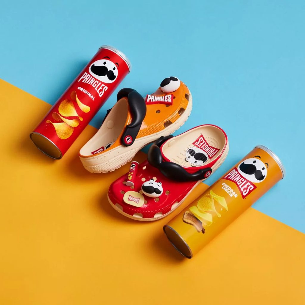 Đôi giày Crocs đượt đặt giữa 2 ống khoai tây Pringles, nằm trên nền cam/xanh