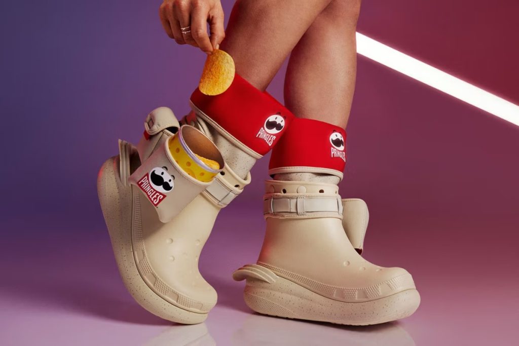 Đôi boot Crocs với thiết kế có hình ảnh Pringles. Một người đang lấy 1 lát khoai tây từ túi đựng