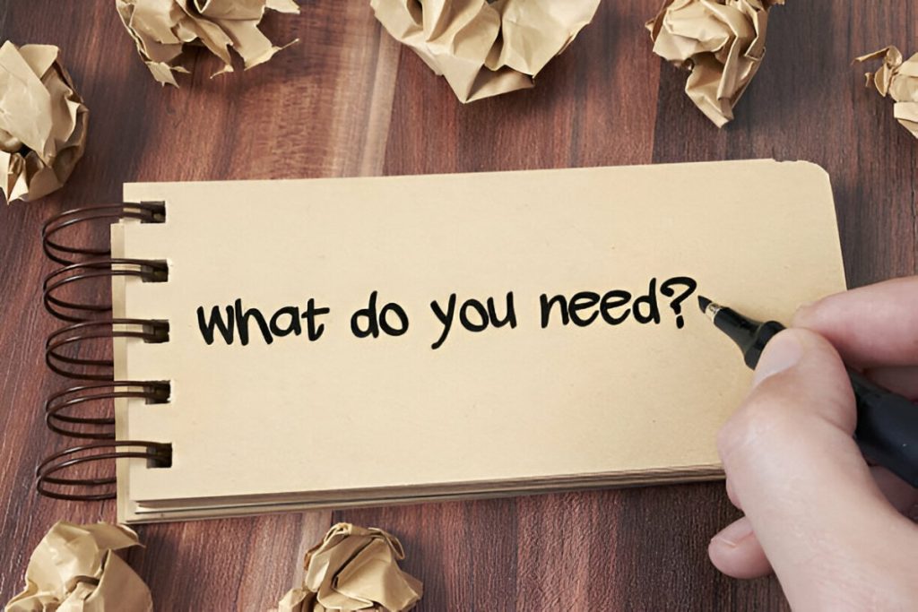 Tập giấy ghi chú với từ "What do you need?" viết mực đen. Tập được đặt trên bàn gỗ với nhiều tờ khác vo cục